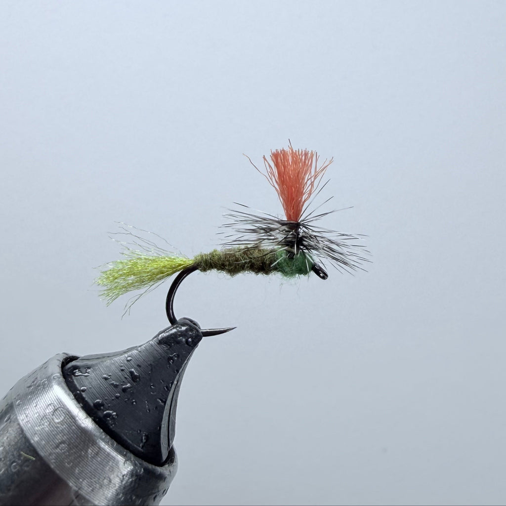 A close-up of a fishing fly tied on a hook, held by a metal clamp against a plain light gray background. The fly has a green body, a bright orange hackle, and light yellow-green hackle hair.